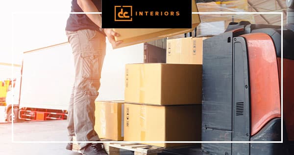 A person stacks cardboard boxes onto machinery in a warehouse. A truck is parked nearby. The image has a "DC Interiors" logo at the top.