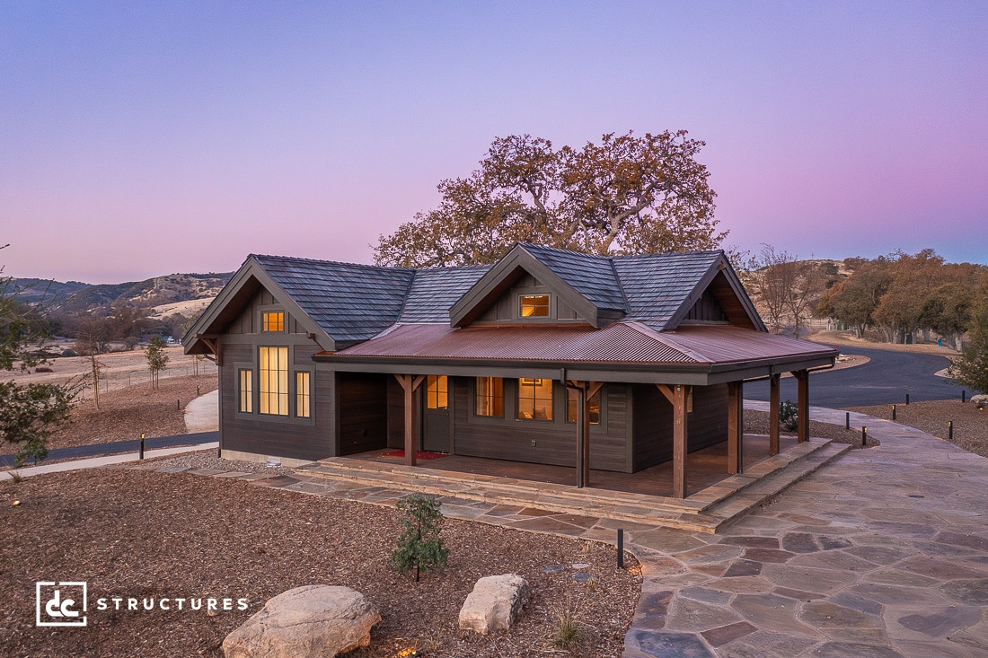 A rustic single-story wooden cabin with a covered porch sits on a stone pathway in a dry open landscape at sunset.
