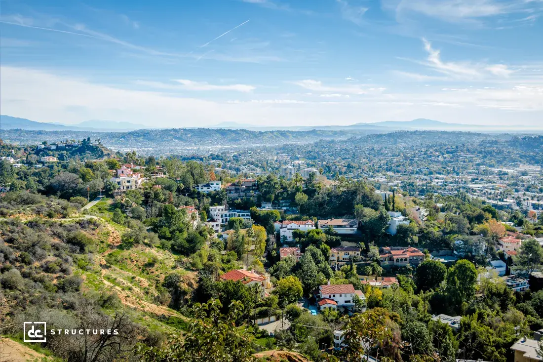 A scenic view overlooking a city with houses among green hills, trees, and clear skies, with mountains in the distant background.