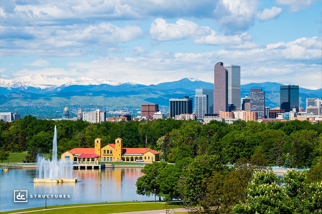 A cityscape of Denver, Colorado with a lake, fountain, yellow building, green trees, skyscrapers, and snow-capped mountains.