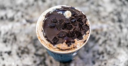 A cup of ice cream topped with chocolate chunks and a drizzle of chocolate syrup sits on a textured gray marble surface, reminiscent of the rustic charm found in apartment barn kits.