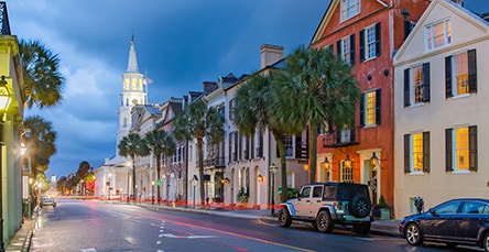 A quaint street scene at dusk with historic buildings lit by streetlights. The buildings vary in color, with some red and others white, set against a dramatic evening sky. Palms line the street, which has light traffic including a parked Jeep. A church steeple is visible in the background beside charming apartment barns.