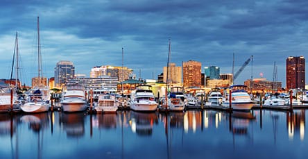 A marina filled with docked boats is seen in the foreground, with the calm, reflective water below and a city skyline illuminated by lights in the background. The sky is overcast, adding a moody ambiance to the scene, while apartment barn kits stand as unique structures blending rustic charm with urban life.