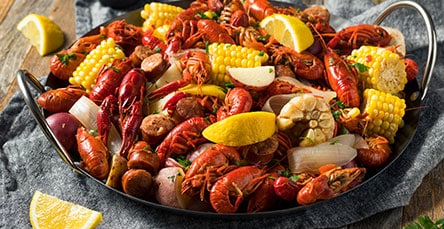 A platter filled with a colorful crawfish boil consisting of seasoned crawfish, sliced sausage, corn on the cob, potatoes, and lemon wedges. The ingredients are garnished with fresh herbs and presented on a rustic wooden table in an apartment barn kit setting.