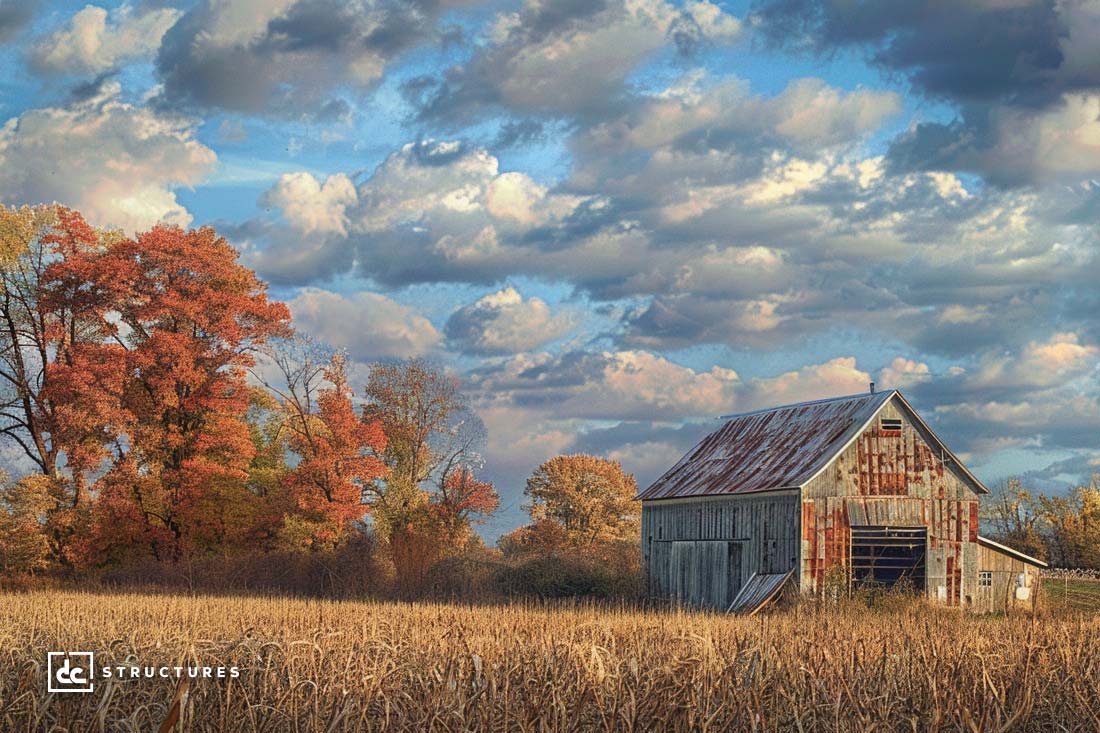 A rustic wooden barn with a rusted roof stands amid a field of tall, golden grasses. In the background, vibrant autumn trees in shades of red, orange, and yellow are set against a sky filled with fluffy clouds. The image evokes a serene, rural atmosphere reminiscent of modern apartment barn kits.