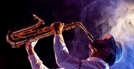 A musician wearing a hat passionately plays a saxophone against a smoky, vibrant background. The scene, reminiscent of the creative energy found in apartment barn kits, is lit with colorful, dramatic lighting that emphasizes the musician's expressive performance.