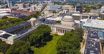 Aerial view of a university campus with large trees and lawns. Prominent in the center is a historic domed building with columns. Surrounding buildings include modern structures, some resembling apartment barn kits. The background features a cityscape with numerous buildings and greenery.