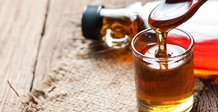 A close-up image of a glass container filled with golden syrup, with syrup being poured into it from a spoon. A glass bottle filled with more syrup lies on its side in the background atop a rustic wooden surface, reminiscent of the charm found in cozy apartment barn kits.