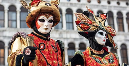 Two individuals dressed in ornate, colorful Venetian costumes and masks stand side by side. The background features a historic building with arched windows, adding to the festive atmosphere reminiscent of detailed apartment barn kits. One person wears a gold and red outfit, the other in white and red.