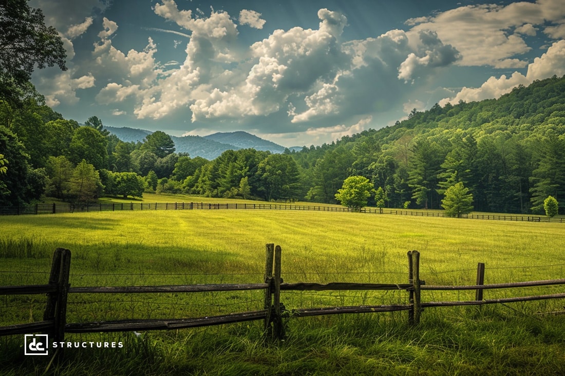 A picturesque landscape featuring a lush green field surrounded by wooden fencing, with a dense forest and mountain range in the background under a partly cloudy sky. The scene is bathed in sunlight, highlighting an apartment barn kit that enhances the natural beauty of the countryside.