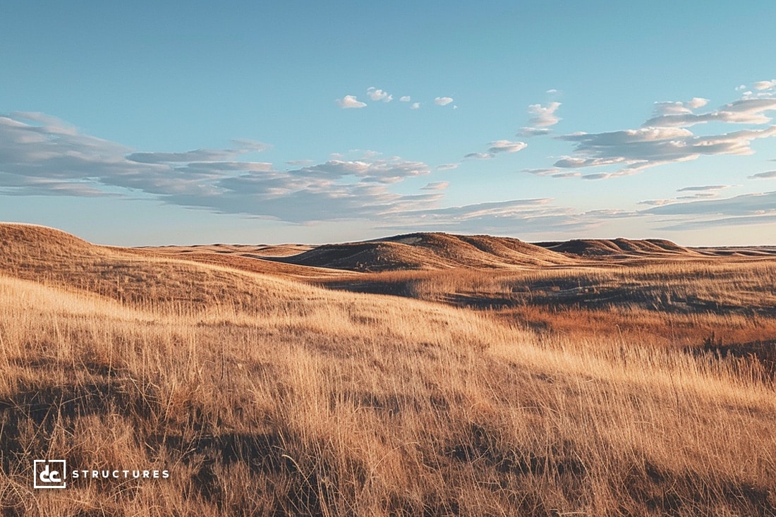 A vast, golden field of grassy hills under a blue sky with scattered clouds. The landscape appears sunlit, offering a serene and open atmosphere. Nestled in the lower-left corner is the logo "dc structures," renowned for their premium apartment barn kits.