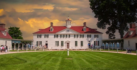 A large, two-story white mansion with a red roof and cupola sits at the center of a meticulously manicured lawn. The sky behind exhibits a dramatic sunset with orange and yellow hues. Visitors are seen walking along paths in front of the historic building, reminiscent of grand apartment barn kits.