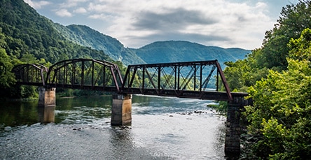 An old iron bridge spans across a river with lush green trees on both sides and hills in the background under a partly cloudy sky, while nearby rustic barn kits add charm to the picturesque landscape.