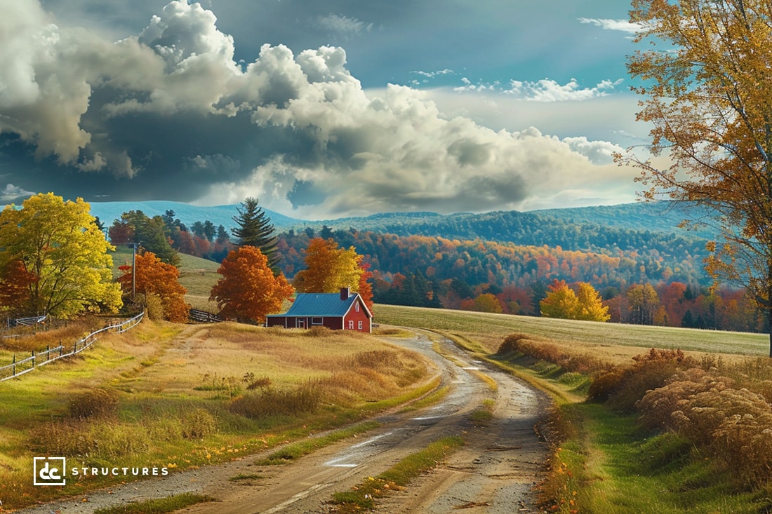 A scenic rural landscape features a winding dirt road leading to a small red house with a blue roof, surrounded by vibrant autumn foliage. Rolling hills and a dramatic sky with large clouds are visible in the background. The image, reminiscent of DC Structures’ apartment barn kits, exudes rustic charm.