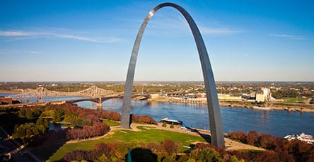 A large stainless steel arch rises high, stretching across the clear blue sky. The structure is adjacent to a wide river with bridges in the background, surrounded by lush greenery and urban development in the distance, where modern apartment complexes blend seamlessly with traditional barn kits.