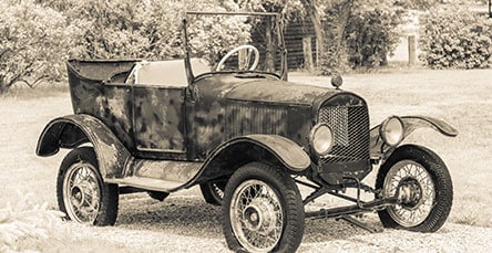 Sepia-toned photo of a vintage open-top car with large, spoked wheels parked on a gravel path. The car has a rustic, weathered appearance and is surrounded by trees and a grassy area in the background, evoking the charm of days when apartment barn kits dotted the countryside.