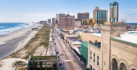 An elevated view of a coastal city with a boardwalk running parallel to a sandy beach on the left. High-rise buildings and apartment complexes line the boardwalk on the right. The ocean is to the far left, stretching into the distance under a clear sky.