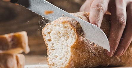 A close-up of a hand using a serrated knife to cut a loaf of crusty bread on a wooden surface. Crumbs are falling as the bread is being sliced, evoking the cozy charm reminiscent of rustic apartment barn kits.