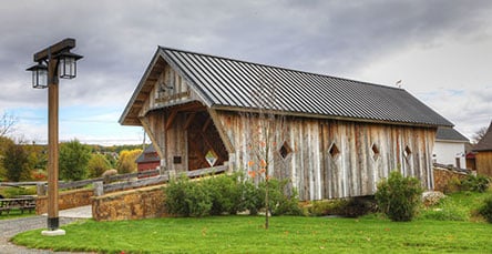 A rustic wooden covered bridge with a black metal roof stands over a small stream. The bridge, reminiscent of designs you might find in apartment barn kits, has diamond-shaped windows and is surrounded by green grass and bushes. A tall wooden lamppost is on the left, and the sky is cloudy.