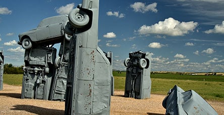 Outdoor art installation featuring several cars partially buried in the ground and stacked vertically, painted grey, resembling a scene crafted from apartment barn kits, set against a background of a grassy field under a blue sky with scattered clouds.