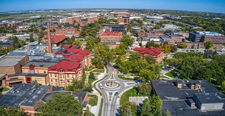 Aerial view of a university campus with multiple red-roofed and brick buildings, green trees, a central roundabout with a fountain, and surrounding urban landscape. Nearby apartment complexes and barn kits speckle the clear, sunny day under the vibrant blue sky.