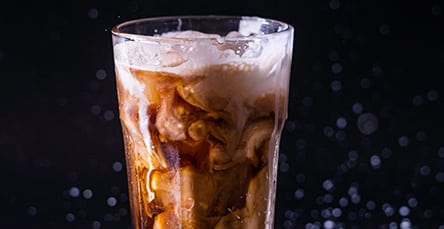 A tall glass filled with iced coffee swirls with cream against a dark, blurred background. The mixture creates a marbled effect as the light catches the frothy top, highlighting the textures of the refreshing beverage—a moment of tranquility before a busy day building apartment barn kits.