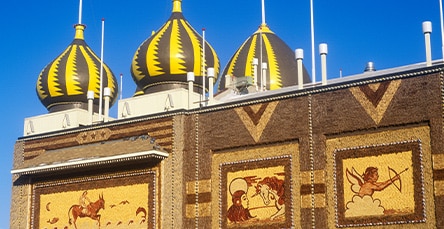 A building with yellow and brown striped domes and detailed mosaics on the walls, resembling intricate apartment designs. The mosaics depict scenes including a rider on a horse and a cherub with a bow and arrow. The sky is clear and blue.