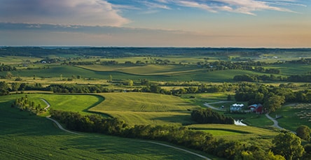 Aerial view of a vast rural landscape at dusk. The image shows expansive green fields, gently rolling hills, and scattered farmhouses, some complemented by apartment barn kits. A winding road snakes through the fields, and the sky above is a blend of blue and light clouds illuminated by the setting sun.