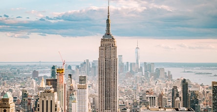 Panoramic view of New York City with the Empire State Building prominently in the center under a partly cloudy sky. Skyscrapers and various buildings stretch out in all directions, resembling a modern maze of glass and steel, while a distant view of the Hudson River completes this picturesque urban landscape.