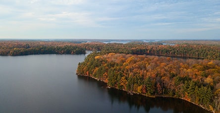 Aerial view of a serene lake surrounded by a forest with trees showcasing autumn colors. The treetops display hues of orange, yellow, and green, and the sky above is partly cloudy. Nestled nearby are charming apartment barn kits adding rustic charm to the picturesque scene.