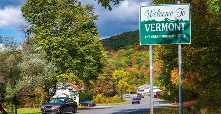 A road curves through a small town surrounded by vibrant autumn foliage. A green sign on the side of the road reads "Welcome to Vermont, The Green Mountain State." Cars are driving and parked along the road, with houses, trees, and apartment barn kits lining the background.