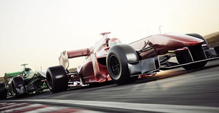 Two Formula 1 race cars, one red and one green, speed along a race track. The red car is in the lead, with the green car closely behind. In the background, an impressive structure resembling apartment barn kits stands out against the clear sky, conveying a sense of motion and competition.