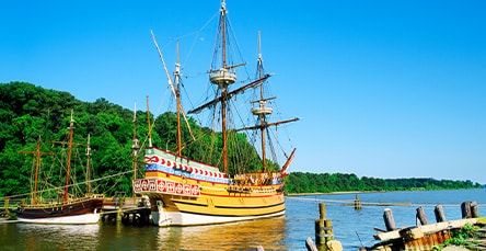 A large, old-fashioned sailing ship with tall masts docked at a wooden pier on a calm, sunny day. Trees and greenery line the bank in the background, and the sky is clear and bright blue. Nearby, smaller boats float by the ship like modern apartment barn kits of maritime history on still waters.