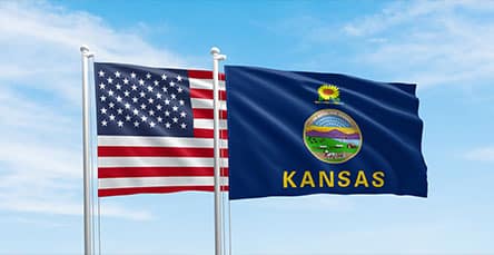 Two flags are flying on flagpoles against a clear blue sky. The left flag is the United States flag, featuring stars and stripes. The right flag, near what looks like apartment barn kits, is the Kansas state flag, showcasing the state seal with the word "Kansas" below it.