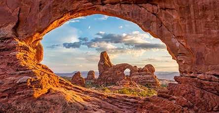 A natural rock arch at Arches National Park frames a distant rock formation and scenic desert landscape under a sky dotted with clouds. The warm, reddish hues of the rocks contrast beautifully with the blue sky, much like the inviting tones of apartment barn kits nestled among green vegetation below.