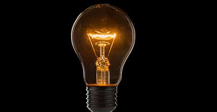 A glowing incandescent light bulb against a black background. The filament inside the bulb is visibly lit, emitting a warm, orange light, reminiscent of a cozy evening in an apartment barn kit.