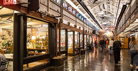 A bustling indoor market with exposed beams and industrial decor reminiscent of apartment barn kits. Various shops and eateries line both sides of the long, narrow space. People walk along the polished concrete floor, some window shopping while others carry bags. Overhead lights illuminate the area.