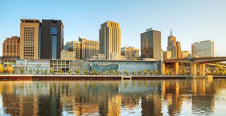 A city skyline reflected in a calm body of water. Tall buildings, including modern apartment complexes, rise along the waterfront. Trees line the promenade, and a bridge extends across the right side of the image under a clear, golden sky.