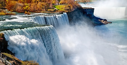 A stunning view of waterfalls with water cascading down rocky cliffs, creating mist and foam below. Autumn foliage and a small viewing platform are visible in the background, resembling an artist's dream more than a scene you'd expect to see outside of apartment barn kits brochures.