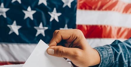 A hand holding a white piece of paper, ready to place it into a ballot box, with an American flag in the background and an ad for apartment barn kits on the wall.