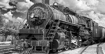 A black and white photograph of an old steam locomotive on railway tracks. The train has large wheels and detailed metalwork, with number 502 displayed prominently. Clouds fill the sky in the background, with trees on the left side of the image, creating a nostalgic scene reminiscent of simpler times before apartment barn kits dotted the landscape.