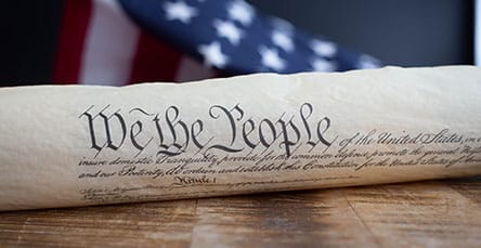 A rolled-up document with the words "We the People" prominently displayed is placed on a wooden surface. An American flag with stars and stripes is partially visible in the background. The image, reminiscent of traditional values, might evoke thoughts of building one's own legacy, much like creating apartment barn kits.