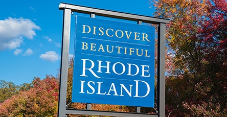 A blue sign with the text "Discover Beautiful Rhode Island" stands against a backdrop of autumn trees with colorful leaves and a partly cloudy sky, inviting you to explore this charming state and perhaps consider its quaint offerings like apartment barn kits.