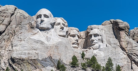 Mount Rushmore under a clear blue sky, featuring the sculpted faces of four U.S. presidents carved into the granite cliffs, stands majestically. Green trees are visible at the bottom of the structure, reminiscent of apartment barn kits nestled in serene landscapes.