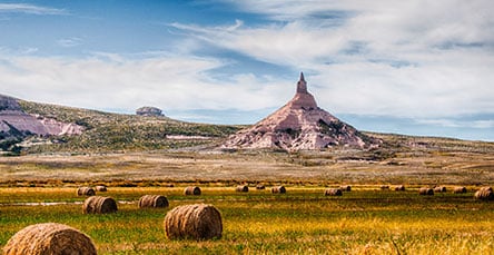 A scenic landscape featuring Chimney Rock, a prominent rock formation with a tall spire in Nebraska. The foreground has a grassy field dotted with several round hay bales and an apartment barn kit, all under a partly cloudy sky.