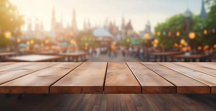 Wooden table in the foreground, against a blurred background of a lively outdoor festival with people, lights, trees, and various structures. The setting appears to be warm and inviting, akin to the cozy charm of an apartment barn kit under a soft evening glow.