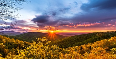 A vibrant sunset casts a golden glow over a mountainous landscape. The sky is filled with dramatic clouds, tinged with hues of orange and purple. Dense forests with autumnal foliage cover the rolling hills and valleys, where quaint apartment barn kits dot the serene and picturesque scene.