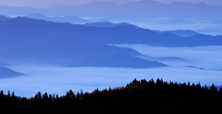 A serene landscape featuring layers of mist-covered blue mountains under a purple sky, with a line of dark forest in the foreground, creating a tranquil, dreamlike atmosphere reminiscent of peaceful retreats once crafted using barn kits.