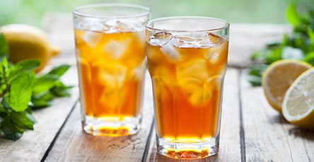 Two glasses of iced tea sit on a wooden table in a cozy nook filled with apartment barn kits. The amber tea is brimming with ice cubes. A partially sliced lemon rests beside the glasses, while fresh mint leaves are scattered around, adding a refreshing touch to the setting.