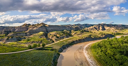 A scenic landscape view shows a winding river bordered by lush greenery on one side and rugged, layered hills on the other. The sky above is partly cloudy with patches of blue peeking through, casting shadows over the hilly terrain. In the distance, an apartment barn kit nestles seamlessly into nature's embrace.